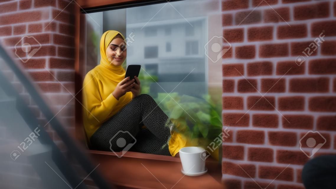 Porträt einer jungen muslimischen Frau, die ein Smartphone benutzt, während sie auf einer Fensterbank in einer gemütlichen Brownstone-Hauswohnung sitzt. Mädchen überprüft Online-Social-Media. Kamerawinkel von