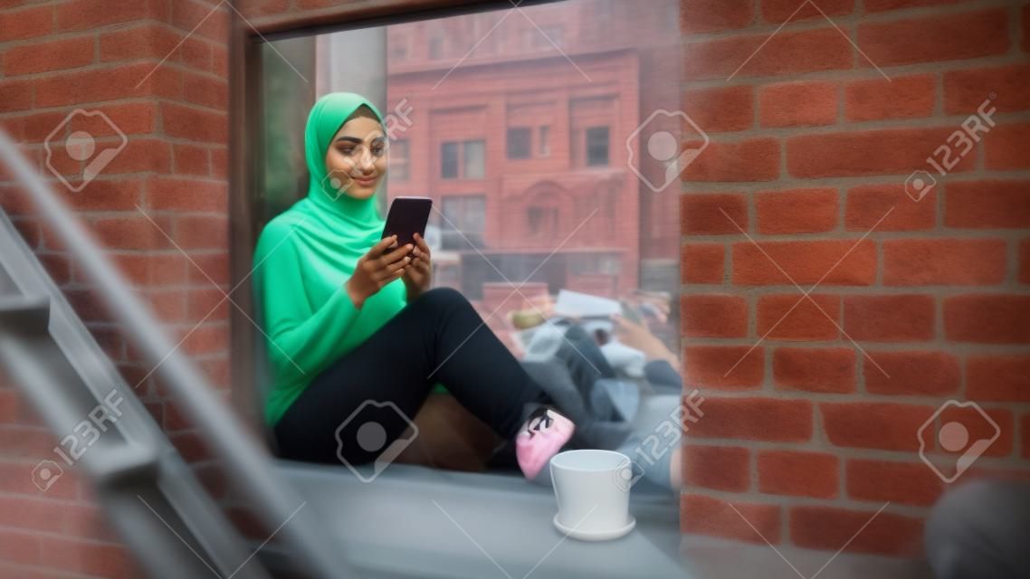 居心地の良いブラウンストーンの家のアパートの窓枠に座りながらスマートフォンを使う若いイスラム教徒女性のポートレート。オンラインソーシャルメディアをチェックする女の子。カメラアングルから
