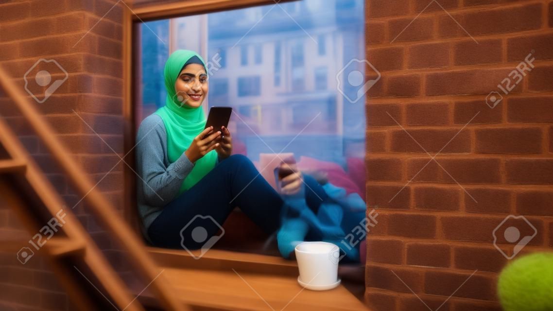 아늑한 브라운스톤 주택 아파트의 창틀에 앉아 스마트폰을 사용하는 젊은 무슬림 여성의 초상화. 온라인 소셜 미디어를 확인하는 소녀. 카메라 각도