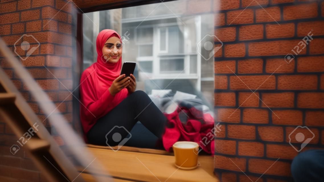 Retrato de uma jovem muçulmana usando um smartphone enquanto está sentada no parapeito de uma janela em um aconchegante apartamento de brownstone. garota verificando mídias sociais on-line. ângulo da câmera de