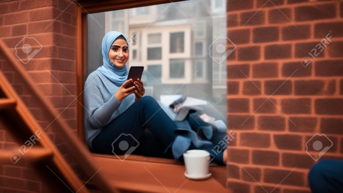 Portret van een jonge moslimvrouw die een smartphone gebruikt terwijl ze op een vensterbank zit in een gezellig bruinstenen huisappartement. meisje dat online sociale media controleert. camerahoek van