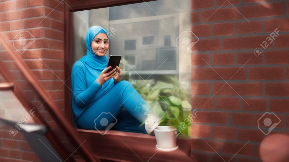 Portret młodej muzułmanki korzystającej ze smartfona, siedzącej na parapecie okna w przytulnym mieszkaniu z piaskowca. dziewczyna sprawdza media społecznościowe online. kąt kamery od