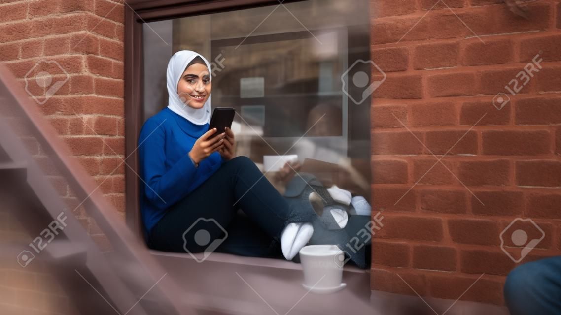Ritratto di una giovane donna musulmana che utilizza uno smartphone mentre è seduta sul davanzale di una finestra in un accogliente appartamento in pietra arenaria. ragazza che controlla i social media online. angolo della telecamera da