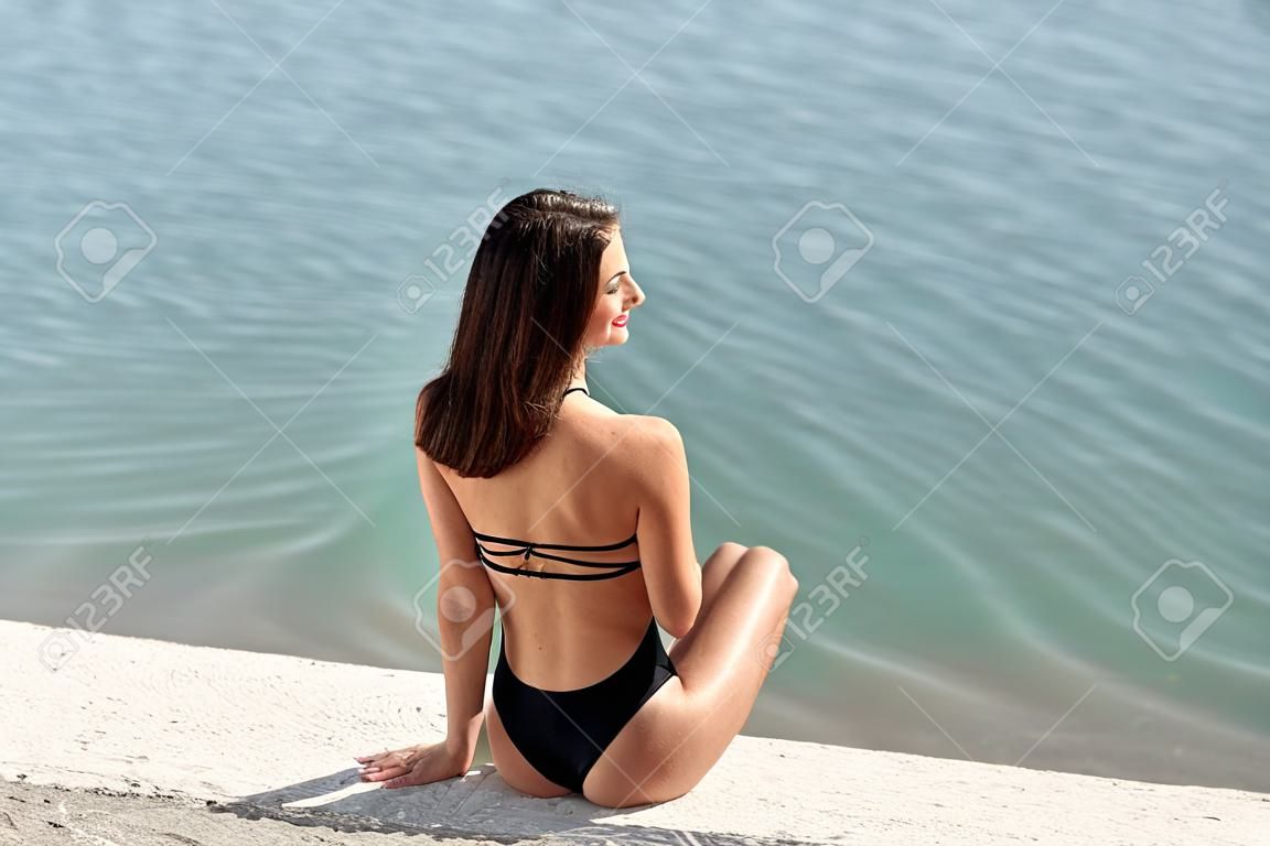 Beautiful young woman with brown hair in black swimsuit on the beach. Hot summer day at the coastline.