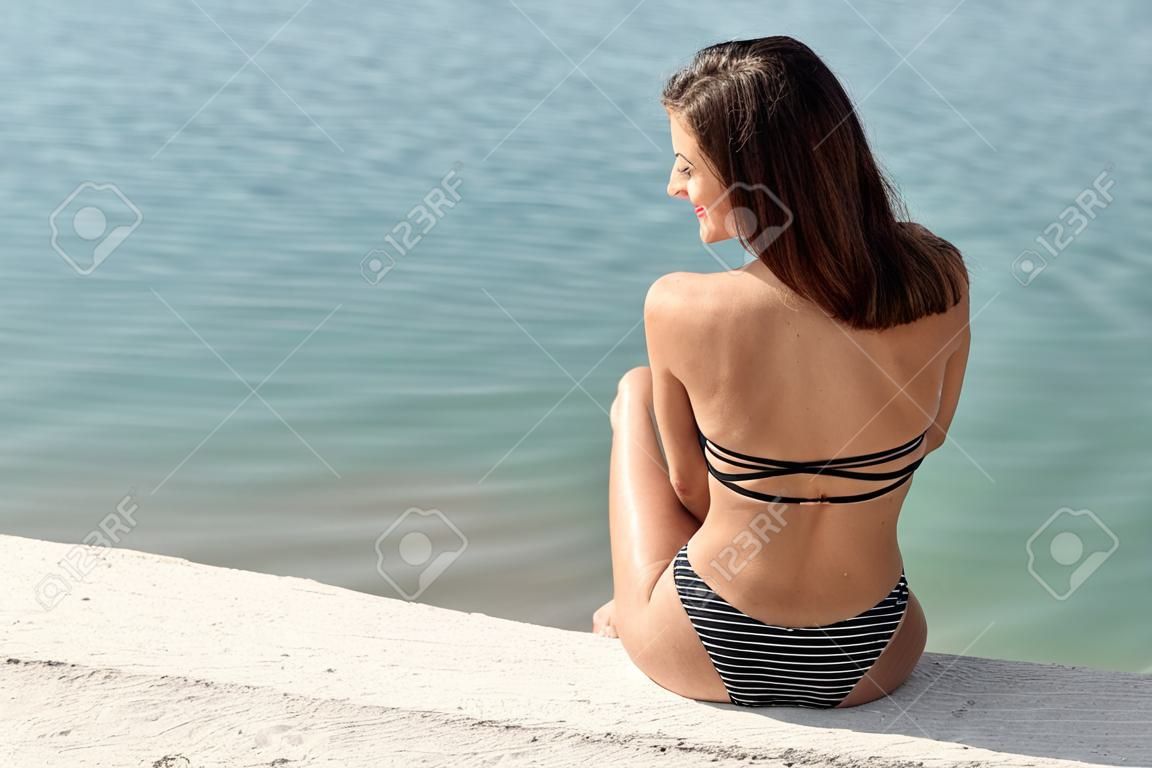 Beautiful young woman with brown hair in black swimsuit on the beach. Hot summer day at the coastline.