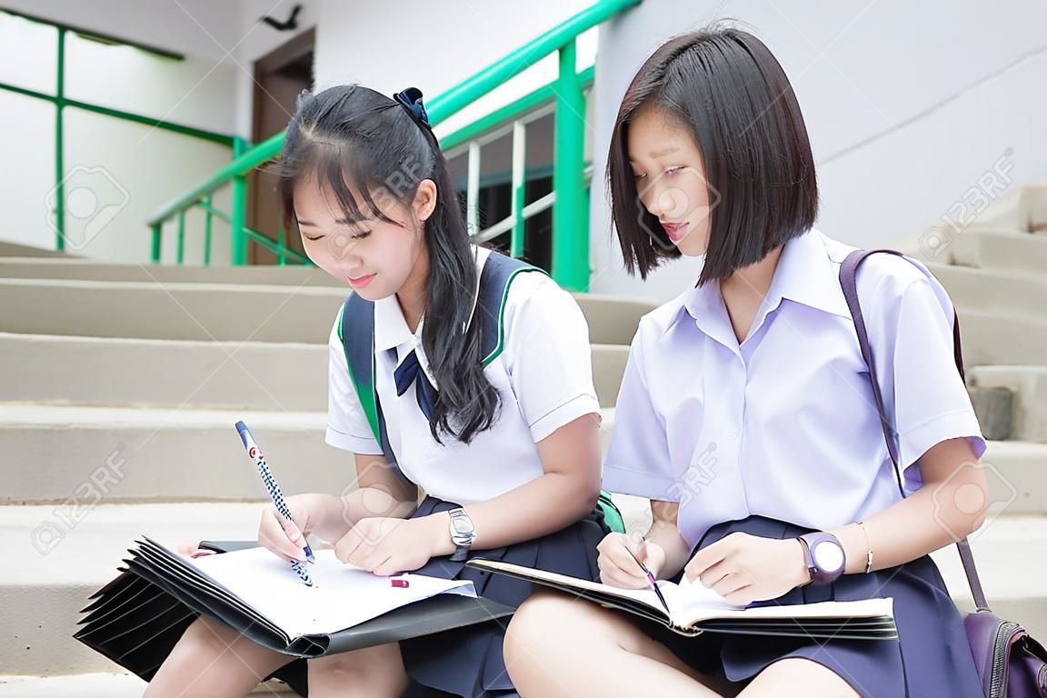 Cute Asian Thai hohe Schülerstudentenpaar Uniform in der Schule auf der Treppe zu diskutieren Hausaufgaben oder Prüfung mit einem glücklichen lächelnden Gesicht zusammen auf einem Gebäude Treppe sitzen