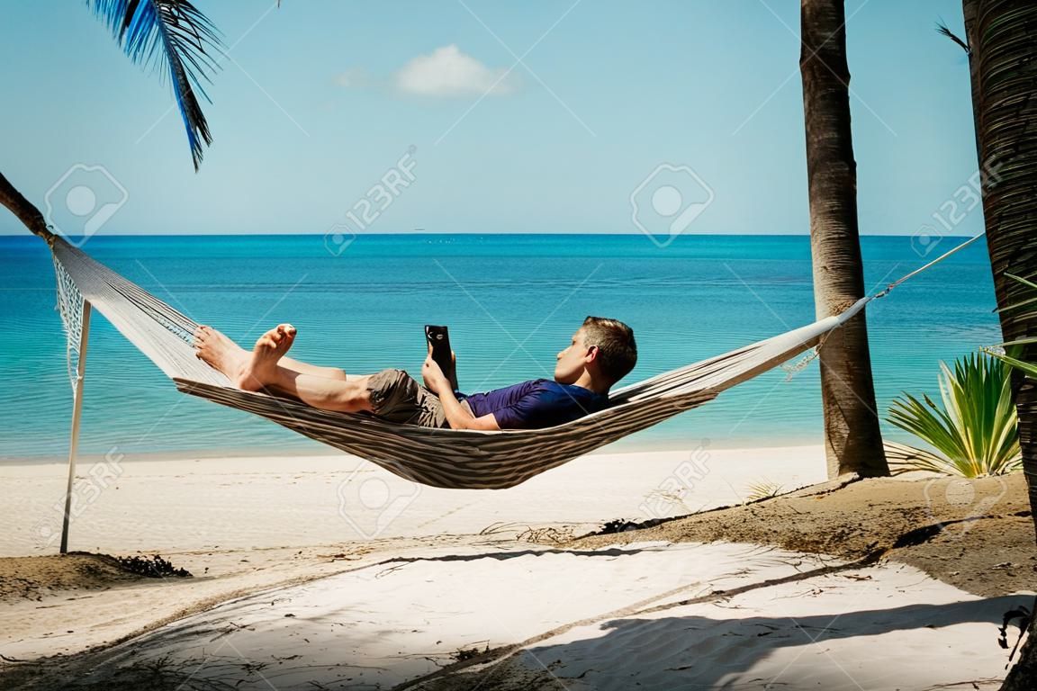 Um jovem relaxa em uma rede na praia enquanto verifica mensagens em seu smartphone.