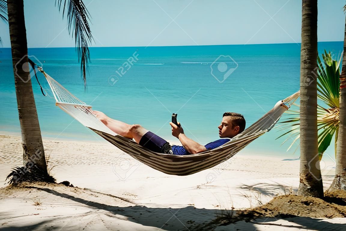 Um jovem relaxa em uma rede na praia enquanto verifica mensagens em seu smartphone.