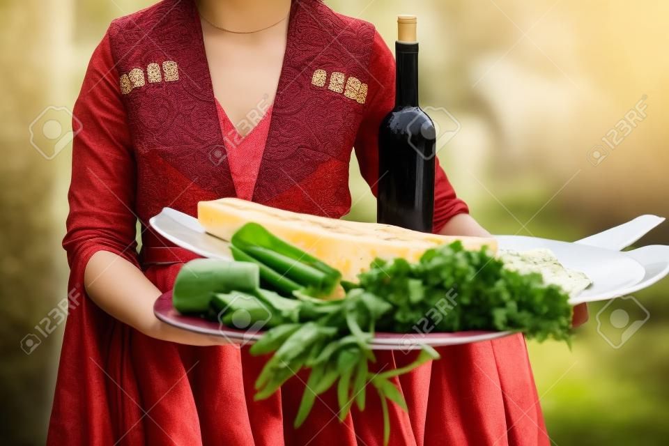 Young beautiful girl wearing traditional georgian dress holds a tray full of traditional georgian food: cheese, bread, greens and wine.