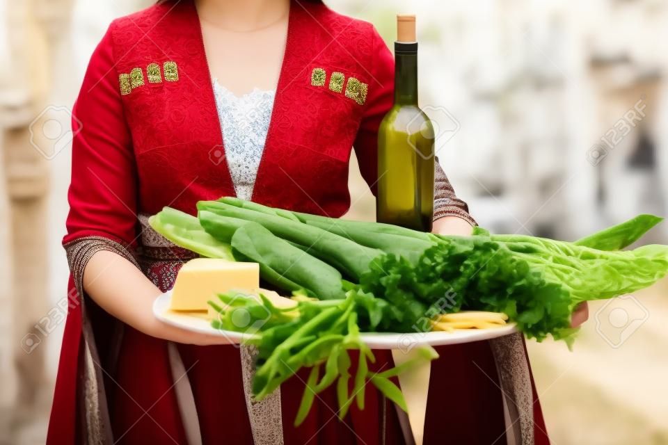 Young beautiful girl wearing traditional georgian dress holds a tray full of traditional georgian food: cheese, bread, greens and wine.