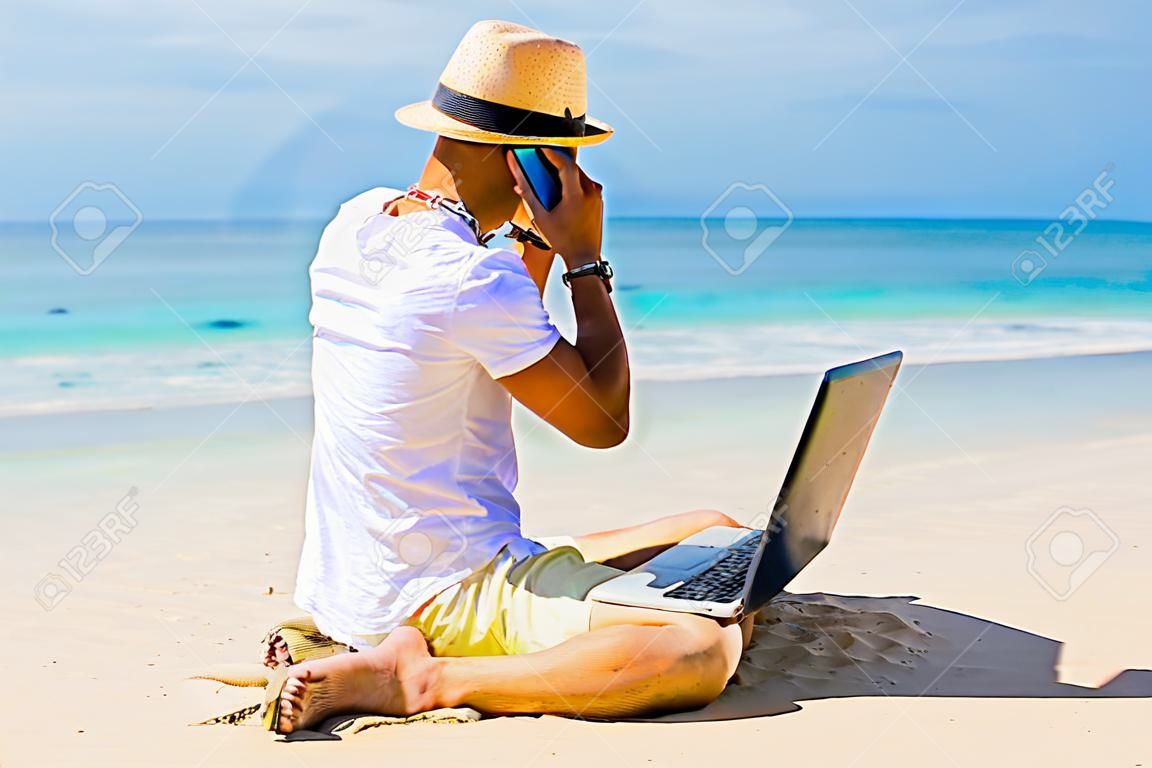 hombre joven en la playa trabajando con ordenador portátil y hablando por teléfono