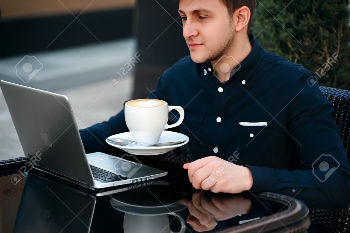 Bel homme boit du cappuchino au café sur la terrasse et regarde dans son ordinateur portable