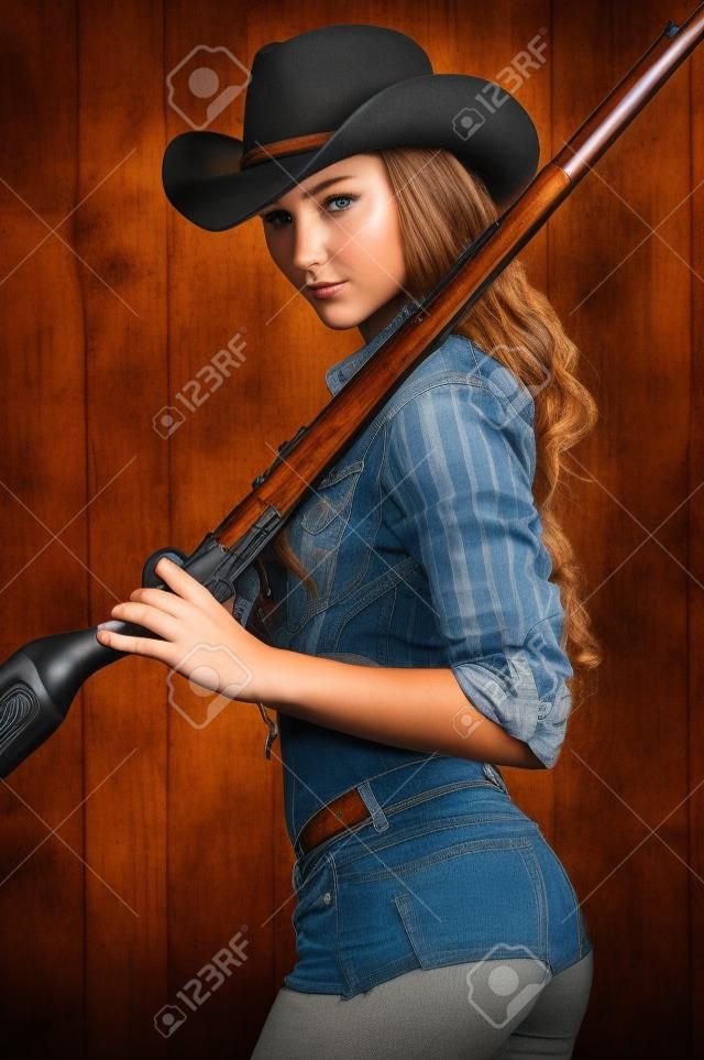 Dangerous beauty. Beautiful young cowgirl carrying gun on her shoulders while standing against the wooden background