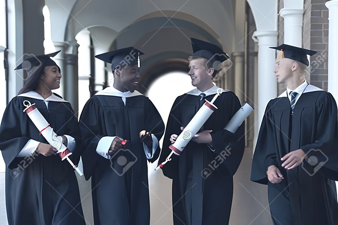 Listo para un futuro brillante. Cuatro graduados universitarios en vestidos de graduación caminando por pasillo de la universidad y hablar