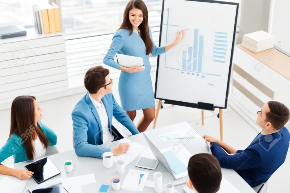 Take a look at this numbers! Top view of beautiful young woman standing near whiteboard and pointing on the chart while her coworkers looking at her and sitting at the table in office