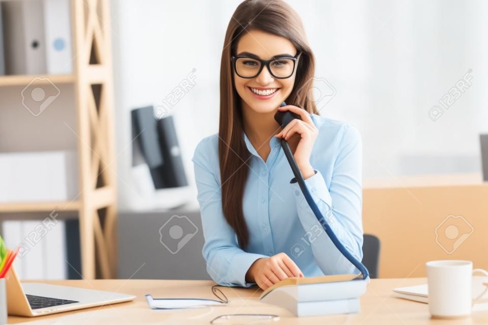 Vi collegherà in un secondo! Allegra giovane bella donna in bicchieri parlando al telefono e guardando la fotocamera con sorriso mentre seduto al suo posto di lavoro