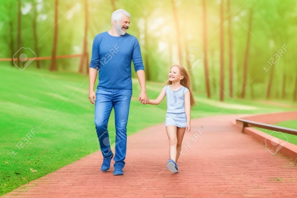 Enjoying great time with father. Front view of cheerful father and daughter holding hands and smiling while walking in park together