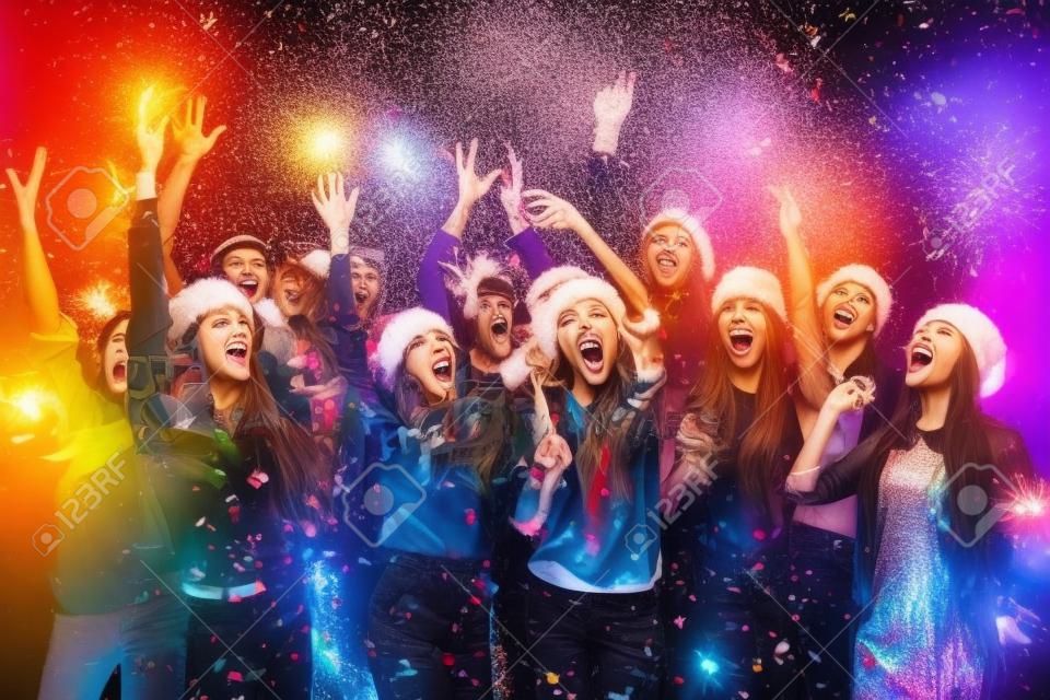 Celebración de Año Nuevo juntos. Grupo de jóvenes hermosas en los sombreros de Santa lanzando confeti de colores y mirando feliz