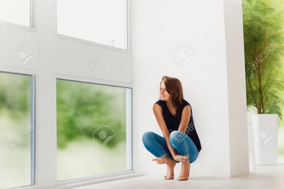 Carefree beauty. Beautiful young woman sitting down while leaning on the wall near the window