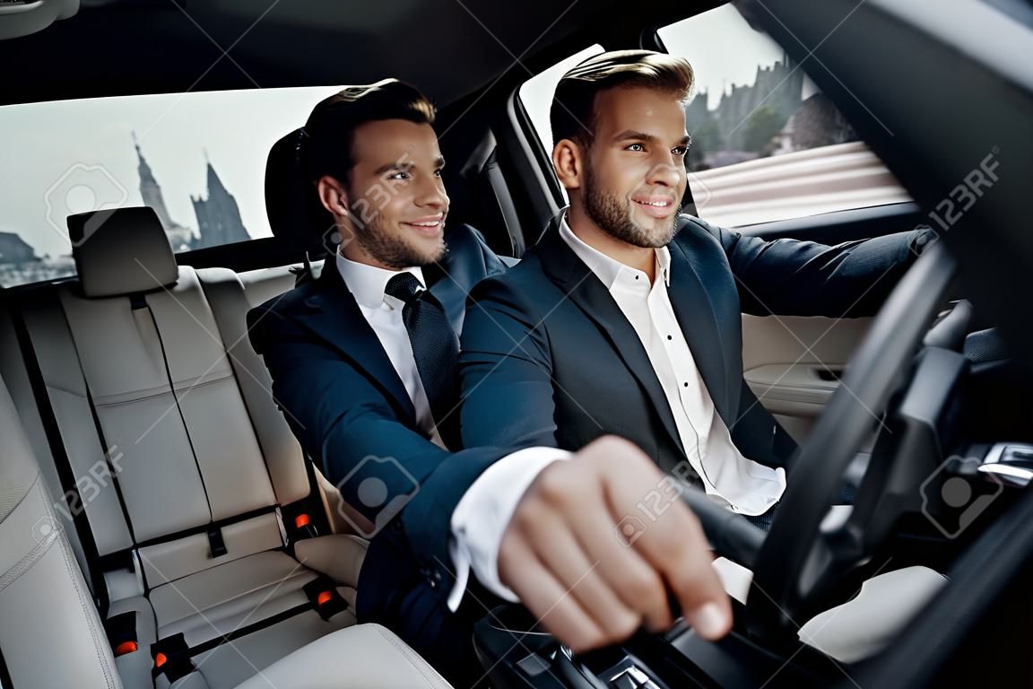 Success in motion. Handsome young man in full suit smiling while driving a car