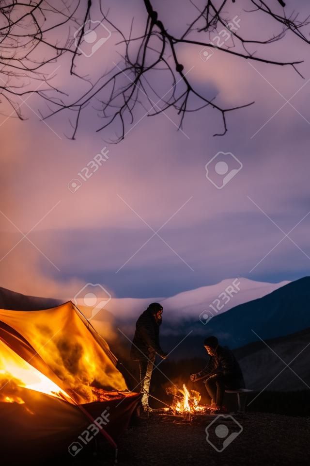 Beautiful young couple warming up by the campfire while camping in the mountains