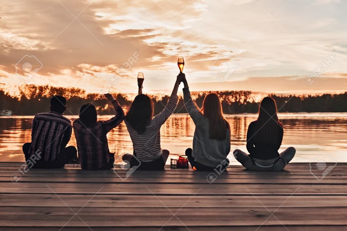 Cheers! Rear view of young woman in casual wear toasting while sitting on the pier among friends