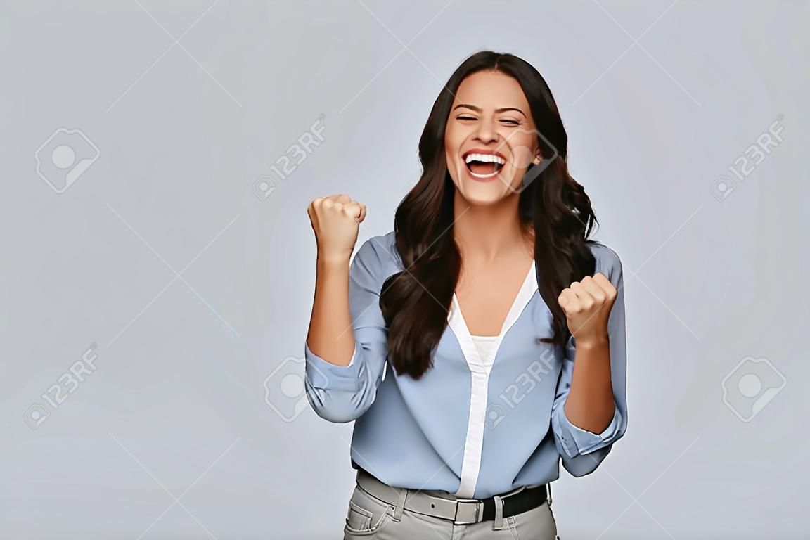 Everyday winner. Attractive young woman punching the air and smiling while standing against grey background