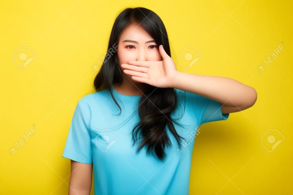 Joven asiática cierra la boca con la mano en un vestido amarillo sobre fondo amarillo