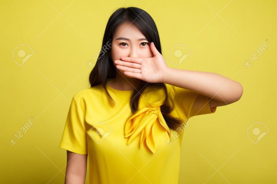 Joven asiática cierra la boca con la mano en un vestido amarillo sobre fondo amarillo