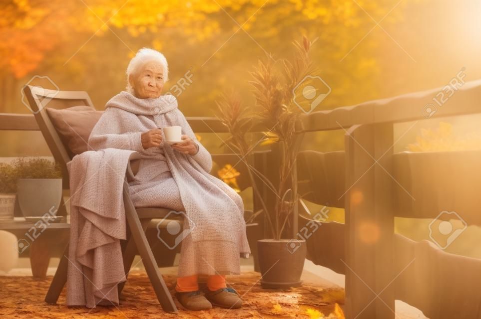 Eine ältere Frau mit einer Tasse, die an einem sonnigen Tag im Herbst draußen auf einer Terrasse sitzt.