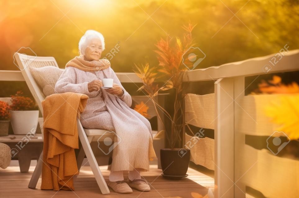 Eine ältere Frau mit einer Tasse, die an einem sonnigen Tag im Herbst draußen auf einer Terrasse sitzt.