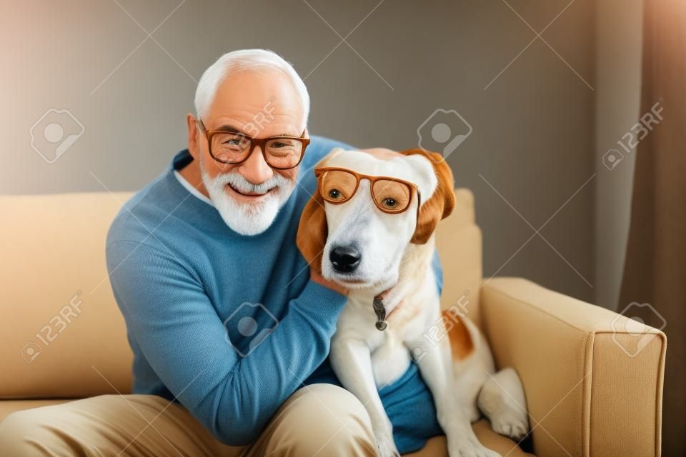 Un hombre mayor sentado en un sofá en el interior con un perro en casa, divirtiéndose.