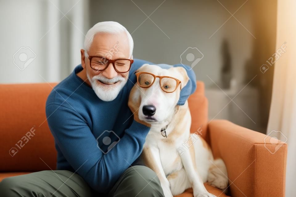 Un hombre mayor sentado en un sofá en el interior con un perro en casa, divirtiéndose.