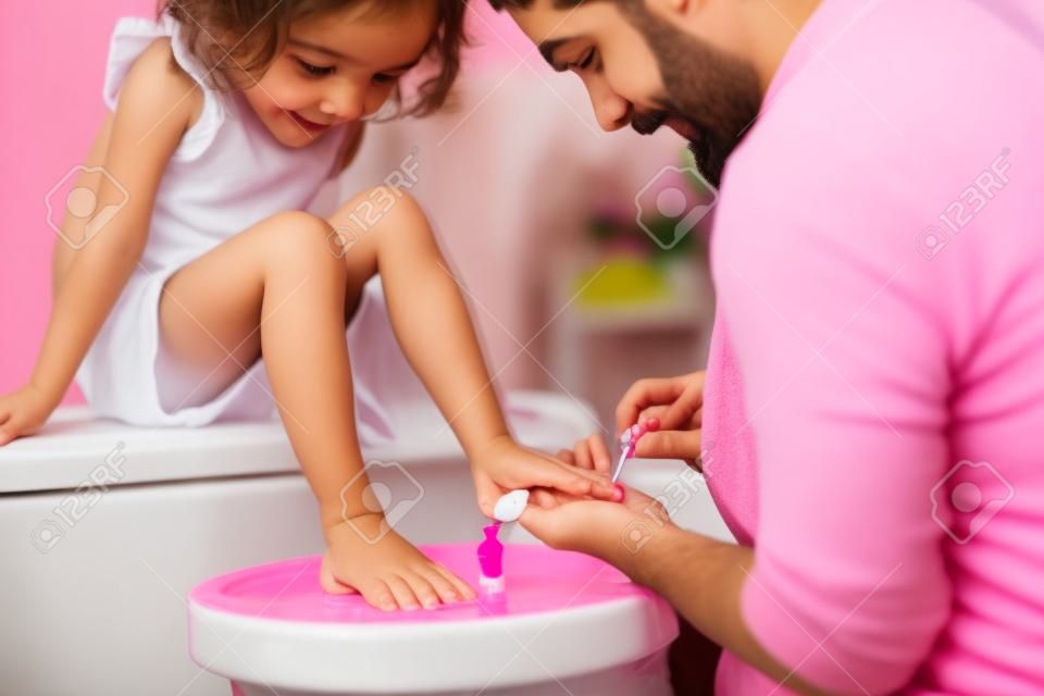 Giovane padre che dipinge le unghie delle piccole figlie in un bagno a casa.