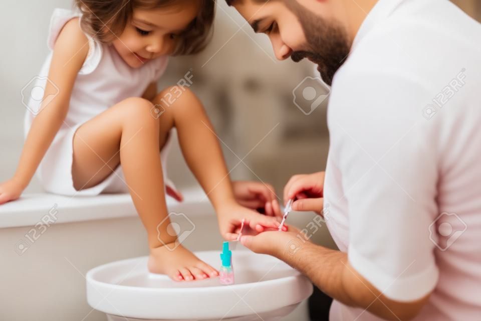 Młody ojciec maluje małe córki paznokcie w łazience w domu.