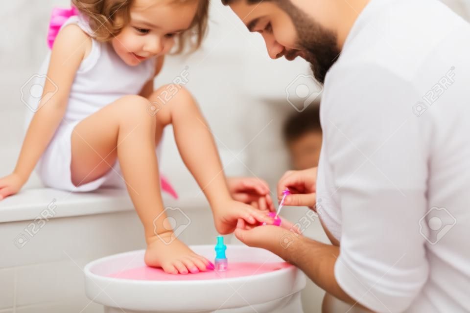 Pai jovem pintando unhas de filhas pequenas em um banheiro em casa.