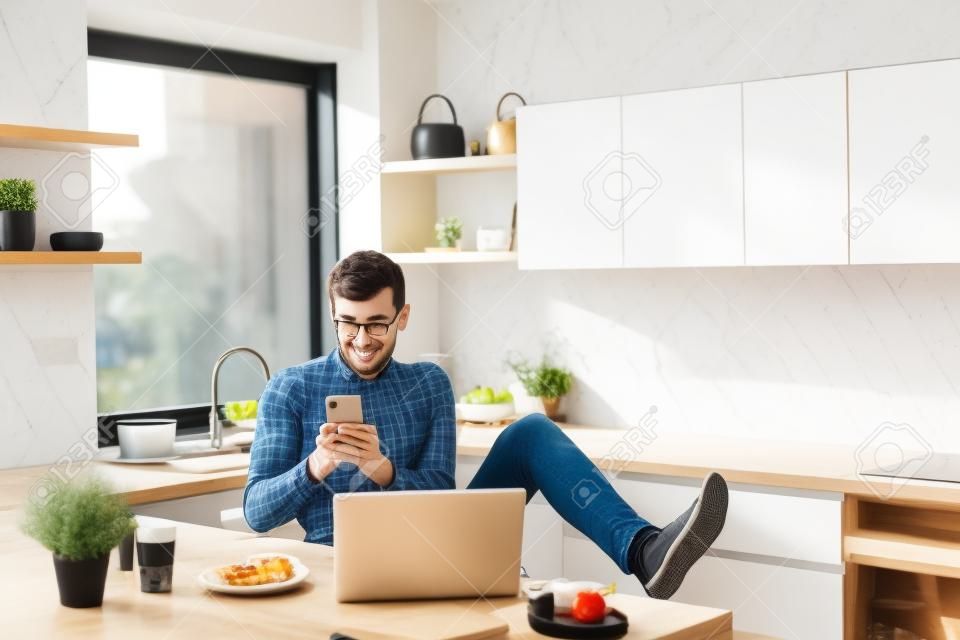 Young man with smartphone and laptop, eating breakfast indoors at home.