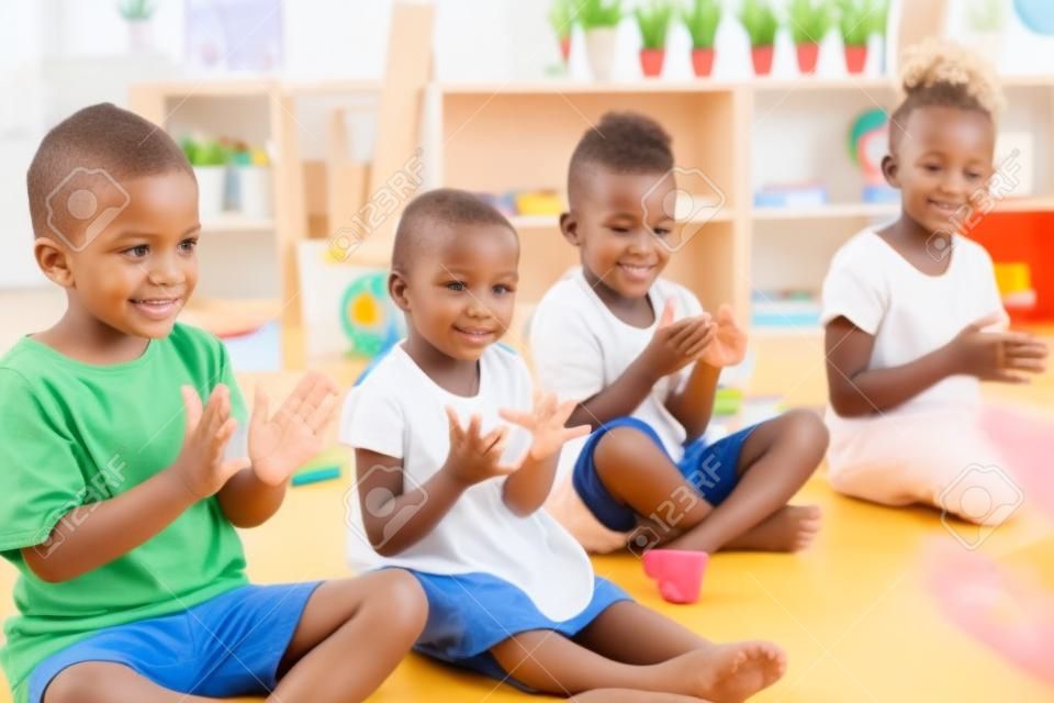 Gruppe kleiner Kindergartenkinder, die drinnen im Klassenzimmer auf dem Boden sitzen und klatschen.