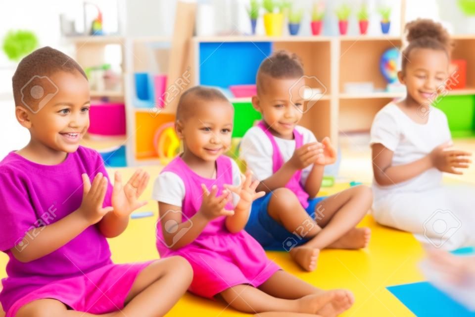 Grupa małych dzieci w wieku przedszkolnym siedzących na podłodze w pomieszczeniu w klasie, klaszczących.