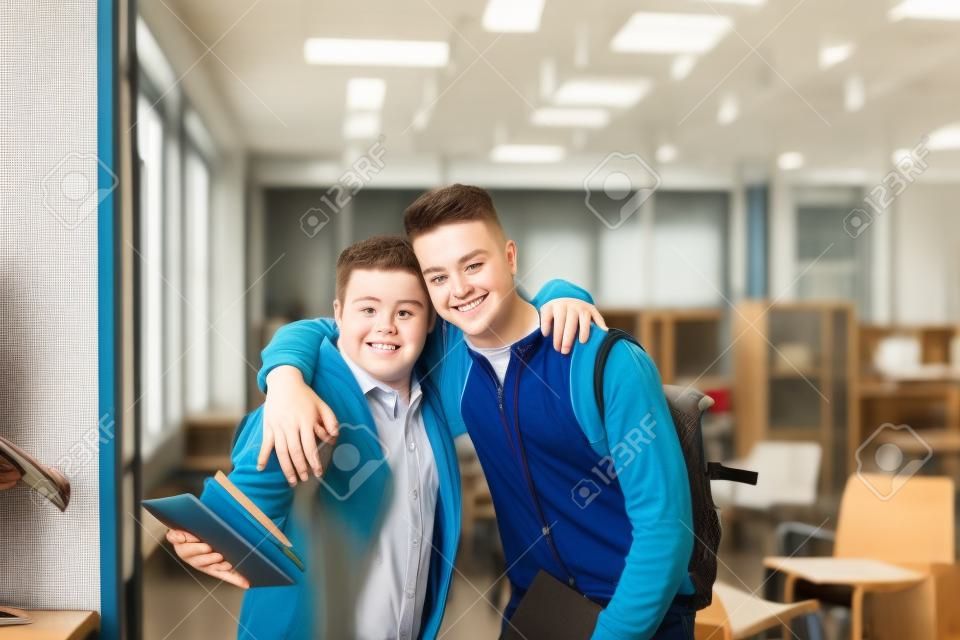 ダウン症の若い男と学校の屋内でカメラを見て周りの腕を持つ彼の家庭教師