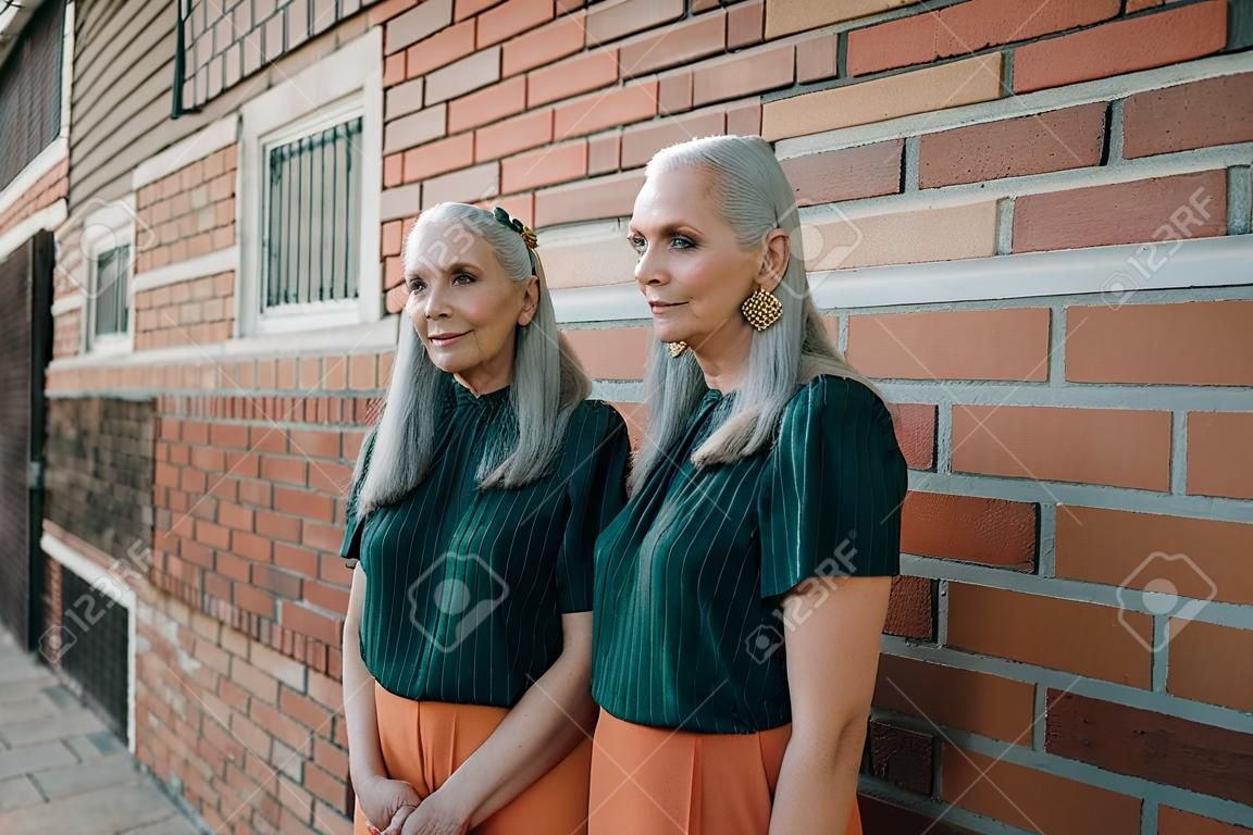 시니어 여성 쌍둥이, 같은 옷을 입고 서서 벽돌 벽 앞에 포즈를 취하고, 도시의 야외.