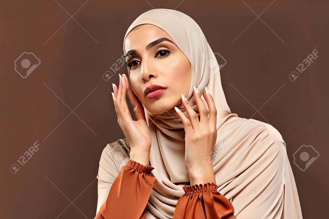Portret pięknej nowoczesnej muzułmanki z naturalnym makijażem ubranej w beżowy hidżab pozowanie na brązowym tle w studio pielęgnacji skóry twarzy kobiece piękno