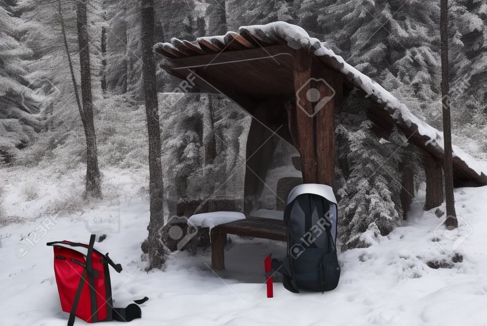 겨울 눈 덮인 숲의 기둥, 나무 껍질 및 가지에서 즉석에서 조급 한 원시적 인 쉼터. 전경에서 빨간색 현대 배낭입니다.