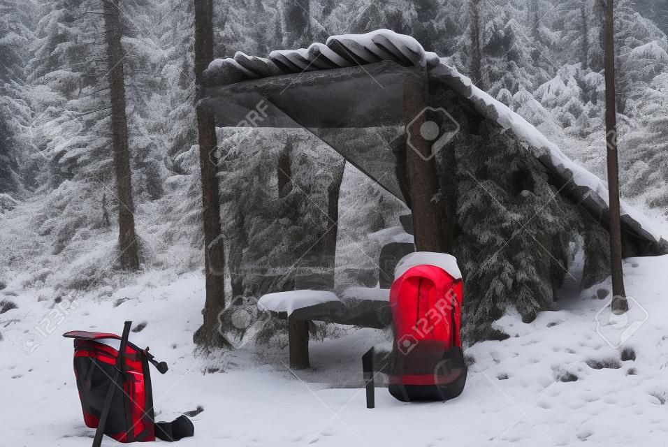 Improvised, deliberately primitive lean-to shelter from poles, bark and branches in the winter snow-covered forest. In the foreground is a red modern backpack.