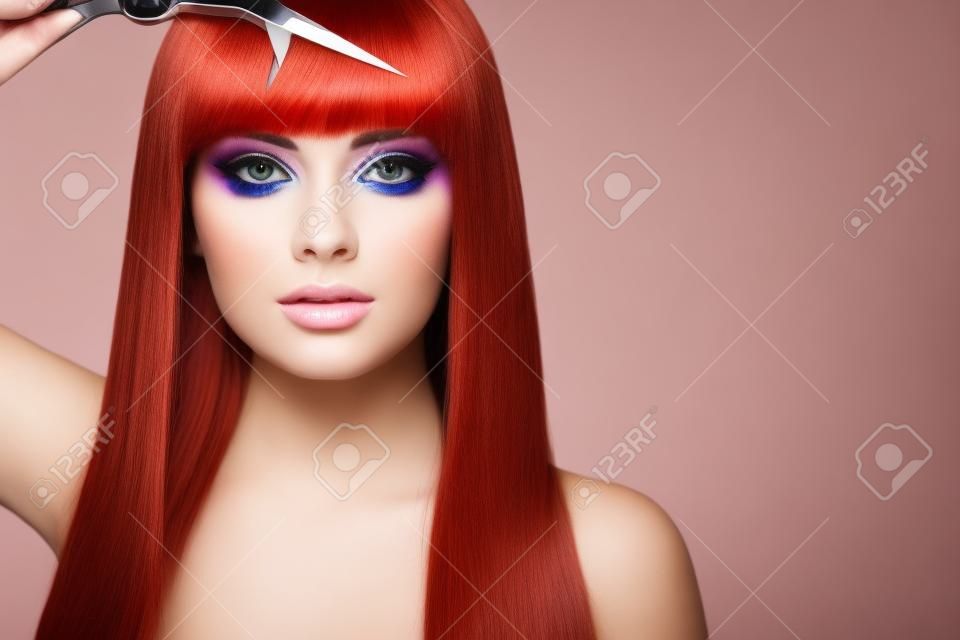 Mulher jovem bonita com uma maquiagem brilhante e um cabelo longo liso segura tesouras de metal. Modelo com cabelo vermelho. Salão de cabeleireiro, corte de cabelo. Cuidados e produtos de cabelo de beleza. Maquiagem perfeita