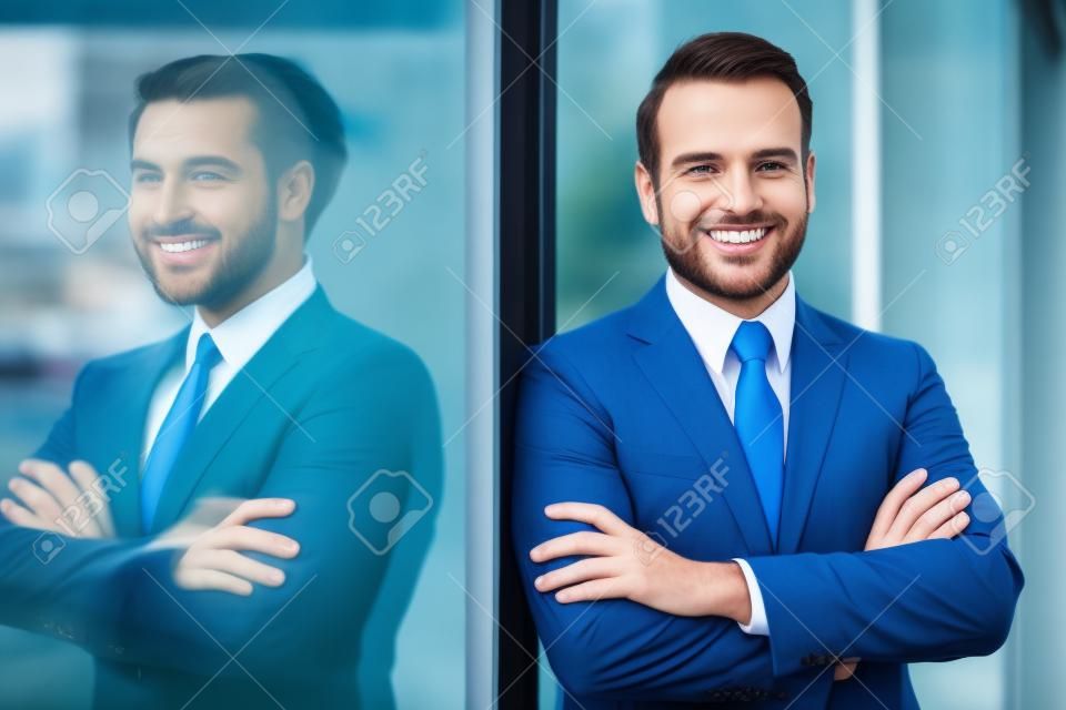 Enkele zelfverzekerde en knappe mannelijke zakenman in blauw pak en stropdas met grijns leunend op raam buiten