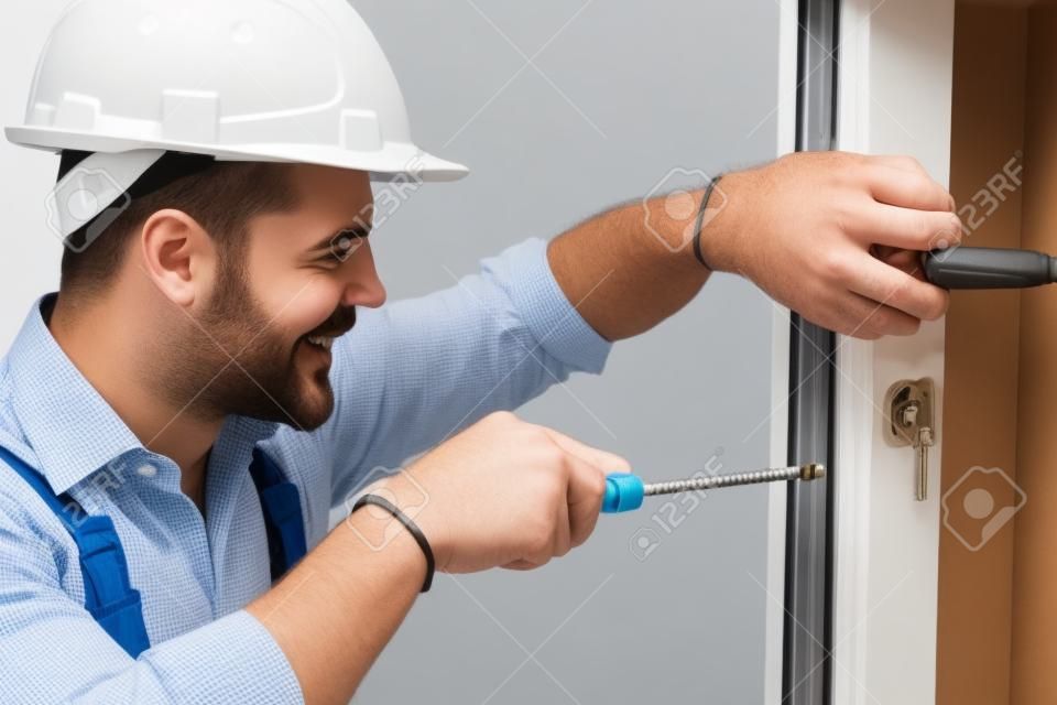 Attractive young builder is installing lock in door. He is holding a screwdriver and kneeling. The man is smiling. He is wearing a helmet
