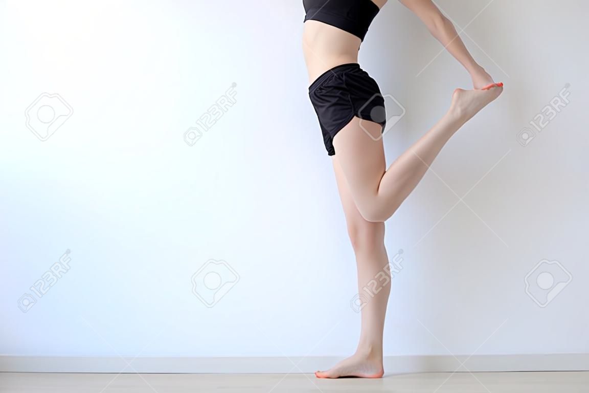 Close up of cheerful slim woman is warming up her body before training. She is standing and raising her leg up. Isolated and copy space in left side