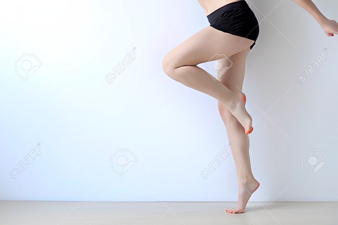 Close up of cheerful slim woman is warming up her body before training. She is standing and raising her leg up. Isolated and copy space in left side