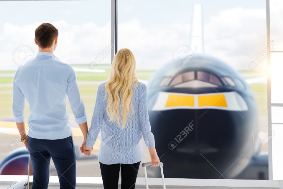 Forward to future. Young loving couple is watching plane though window before departure. They are standing at airport and holding hands